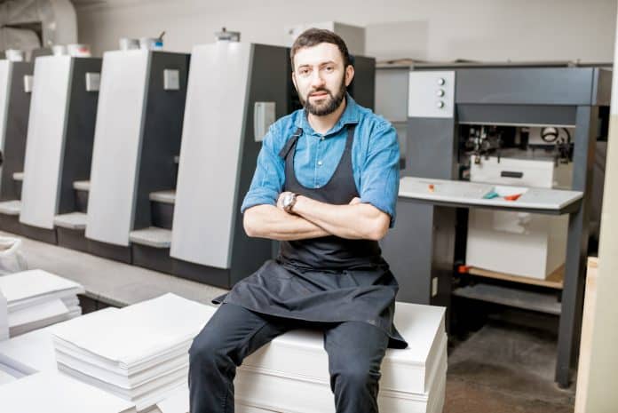 Printer sitting on a stack of papers in front of an offset printing press