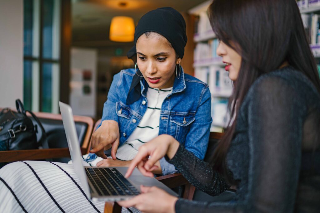 Two women in a library helping each other on a laptop.