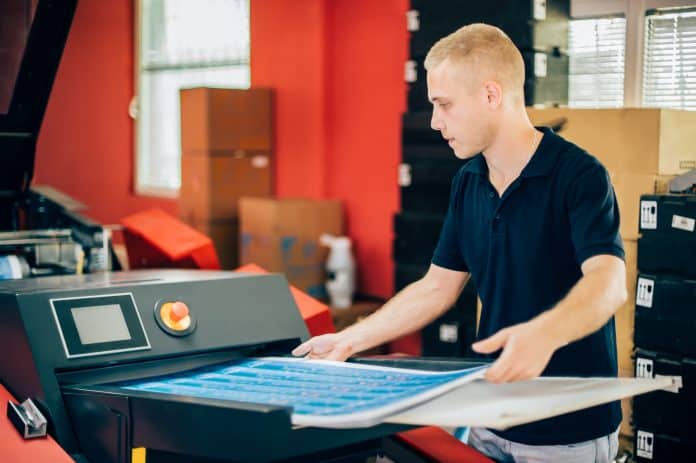 Young man working in printing factory. Man is grabbing prints from the digital printing press